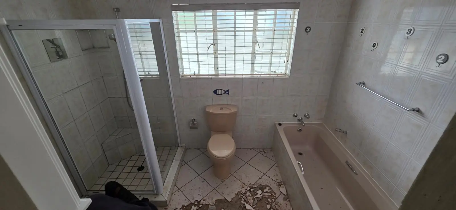 A dated bathroom with cream patterned wall tiles, a built-in bath, a framed shower cubicle and a security-barred window, with loose plaster over the floor mid strip-out.