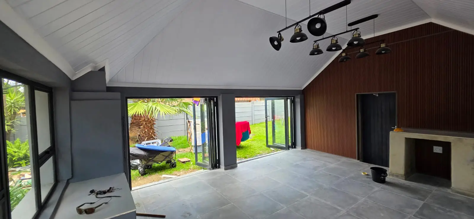 The same space finished as an enclosed entertainment room, with a pitched white ceiling, black industrial track lighting, charcoal aluminium folding doors standing open onto a lawn, a timber-clad feature wall and a built-in braai.