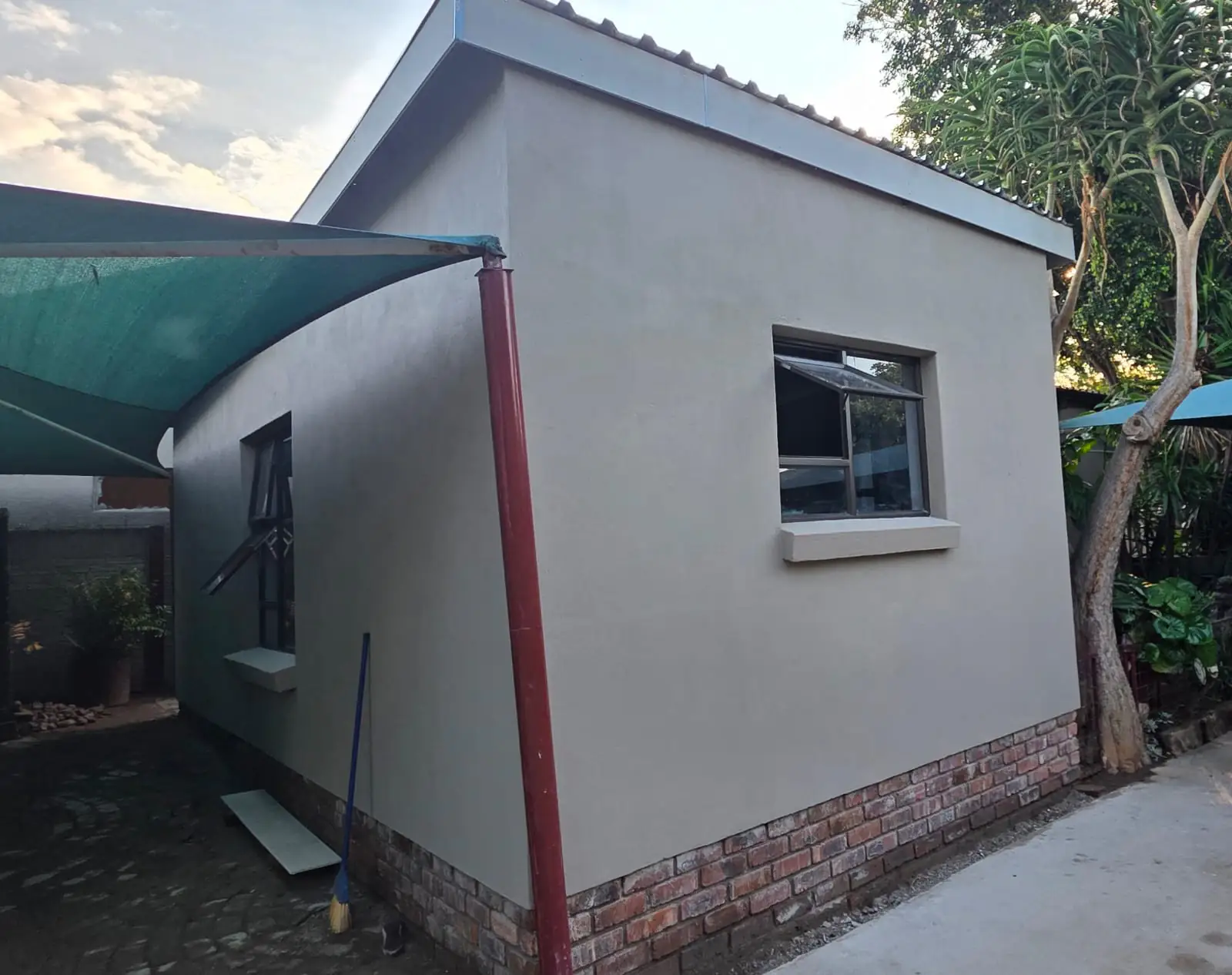The finished outbuilding with smooth plastered walls on a face-brick plinth, a charcoal aluminium window, a boxed roof edge and a new red downpipe, standing on a swept concrete apron.