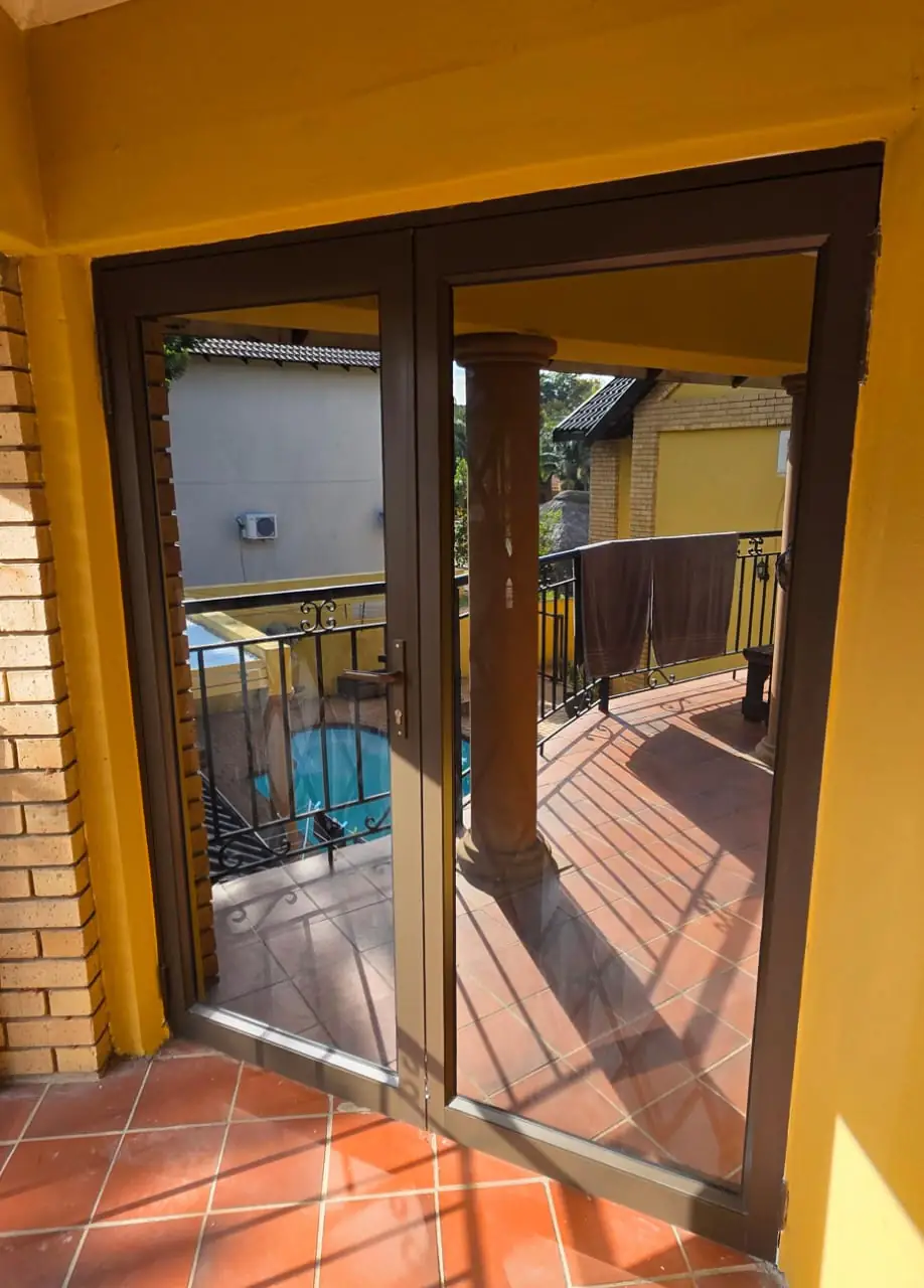 New bronze aluminium double doors with full-height clear glazing in the same opening, looking out past a plastered column to the balcony railing and the pool below.