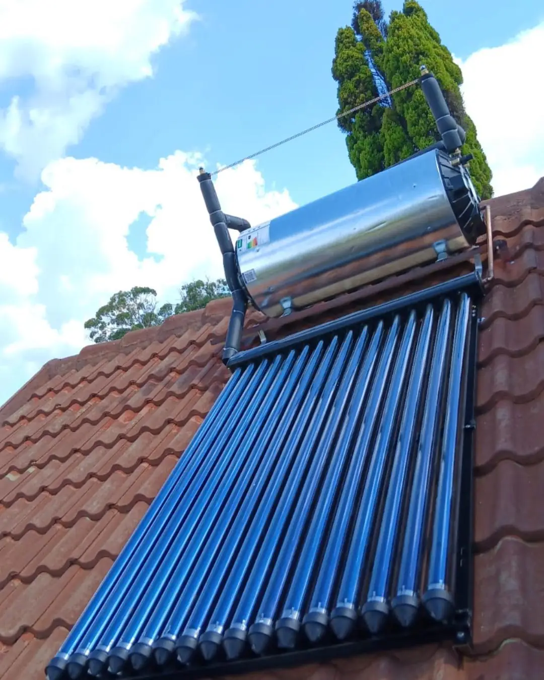 A stainless steel solar geyser cylinder and a bank of blue evacuated tube collectors mounted on a red tiled roof, with copper pipework running down into the roof and a bright cloudy sky behind.