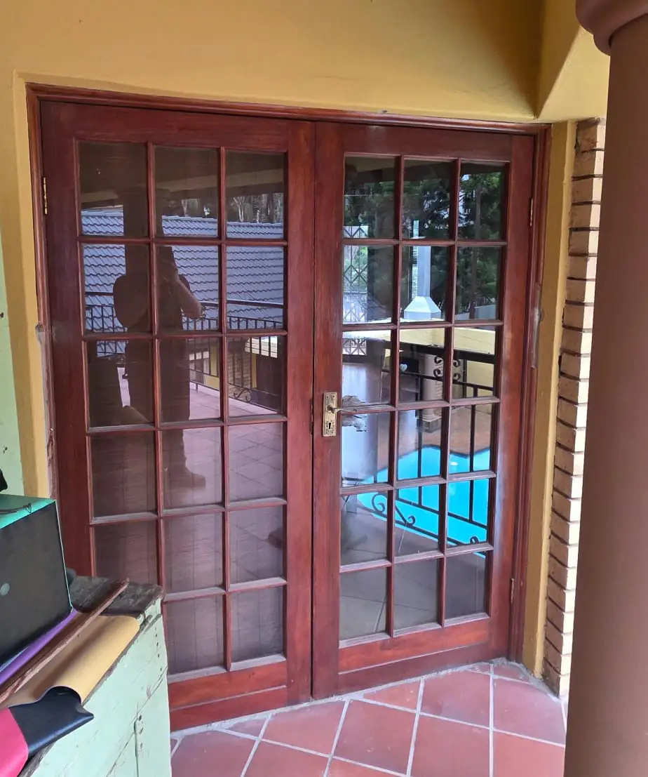 Varnished timber French doors with a grid of small glass panes, leading from a storeroom onto a covered patio with terracotta tiles.